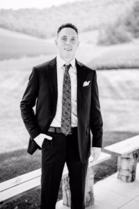 A man in a suit and patterned tie stands outdoors near benches at a Horstmann Homestead wedding, with hills and trees in the background.