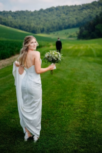 A woman in a white wedding dress holding a bouquet stands on a grassy field at Horstmann Homestead wedding, looking back, while a man in a dark suit walks away in the distance.