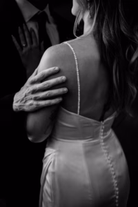 A person in a suit gently holds the shoulder of someone in a buttoned satin dress, shown from the back in black and white at a Horstmann Homestead wedding.