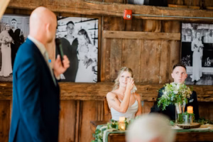 A bride and groom sit at a wooden table decorated with flowers, looking toward a man giving a speech at their Horstmann Homestead wedding in a rustic venue with wooden walls.