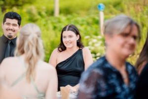 A woman in a black one-shoulder dress sits outdoors at a Horstmann Homestead wedding, surrounded by other guests with lush greenery in the background.