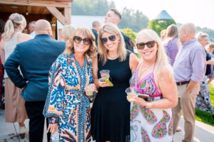 Three women in sunglasses and colorful dresses stand together outdoors at a Horstmann Homestead wedding, smiling and holding drinks, with a group of people mingling in the background.