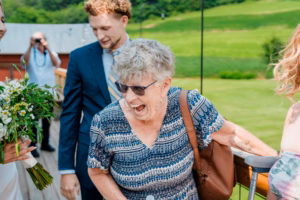 An elderly woman wearing sunglasses laughs while standing outdoors at a Horstmann Homestead wedding, surrounded by formally dressed guests and lush greenery in the background.