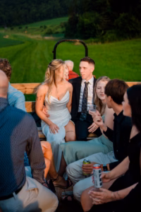 A group of young adults sits together on wooden benches in an open trailer at a Horstmann Homestead wedding, green fields and trees in the background. Some are holding drinks and talking.