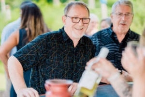 Two men in patterned shirts stand at an outdoor table with drinks during a Horstmann Homestead wedding, one smiling at the camera while others interact in the background.