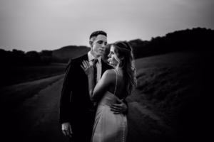 A man in a suit and a woman in a formal dress stand close together on a rural road at the picturesque Horstmann Homestead wedding, with trees and hills in the background.