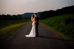 A bride and groom stand embracing on a quiet country road at sunset, surrounded by green fields and trees, capturing the romance of a Horstmann Homestead wedding.