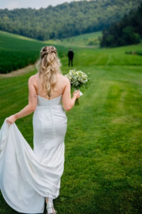 A bride in a white dress holding a bouquet walks across the green lawn at Horstmann Homestead wedding venue, heading toward a person in the distance, framed by hills and trees in the background.